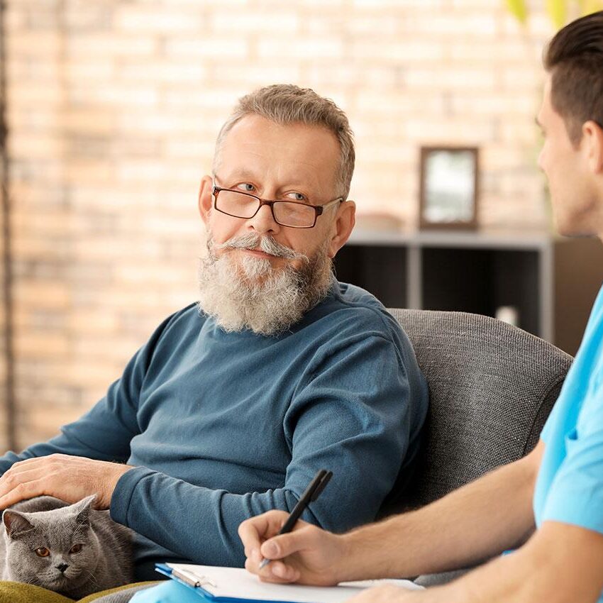 An Elmes Homecare carer sitting with an older man at home, taking notes to understand his care needs